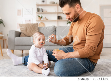 Father Feeding Little Baby Daughter Sitting On Floor At Home 70931189