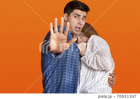 Do not come closer. Frightened female hiding at man chest, guy is terrified, makes stop sign with his hand Do not come closer. Frightened female hiding at man chest, guy is terrified, makes stop sign with his hand 70931307