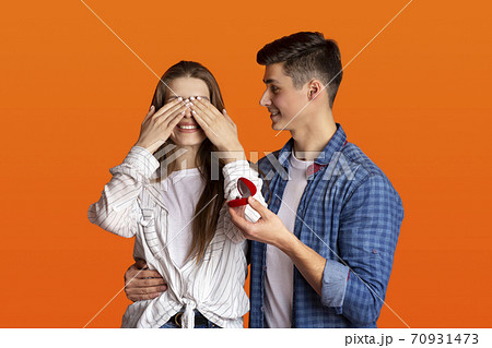 Happy man makes surprise and gives ring in red box, woman covers her eyes with hands Happy man makes surprise and gives ring in red box, woman covers her eyes with hands 70931473