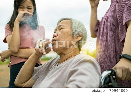Asian senior woman holding a cigarette smoking,smokers elderly smoking near people in family,daughter, granddaughter closing nose,pollution from cigarettes smoke,dangerous to health,addiction,life Asian senior woman holding a cigarette smoking,smokers elderly smoking near people in family,daughter, granddaughter closing nose,pollution from cigarettes smoke,dangerous to health,addiction,life 70944899