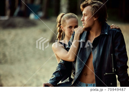 young couple of bikers closely looking at each other on black motorcycle on road near green forest young couple of bikers closely looking at each other on black motorcycle on road near green forest 70949487