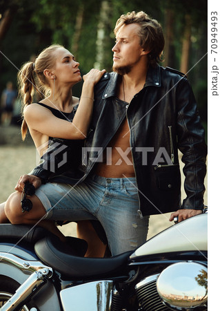 young couple of bikers closely looking at each other on black motorcycle on road near green forest young couple of bikers closely looking at each other on black motorcycle on road near green forest 70949493