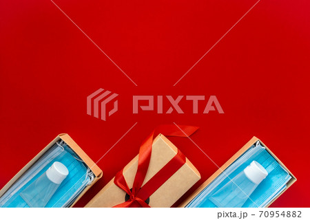 Packing a Christmas present during Coronavirus epidemic. Face masks and hydroalcoholic gels inside a cardboard box decorated with a bow on a red background. Packing a Christmas present during Coronavirus epidemic. Face masks and hydroalcoholic gels inside a cardboard box decorated with a bow on a red background. 70954882