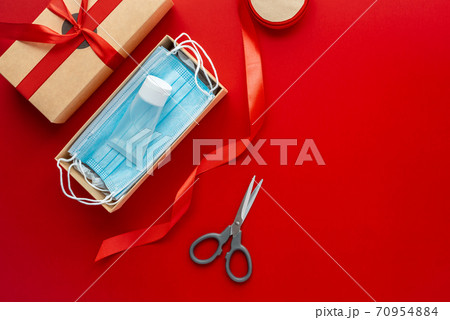 Packing a Christmas present during Coronavirus epidemic. Face masks and hydroalcoholic gel inside a cardboard box decorated with a bow on a red background. Packing a Christmas present during Coronavirus epidemic. Face masks and hydroalcoholic gel inside a cardboard box decorated with a bow on a red background. 70954884