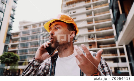 Angry male architect in orange hard hat talks swears on phone at construction site. Builder in plaid shirt emotionally displeased negotiates by cell phone. Screaming stressed architectural designer 70964413