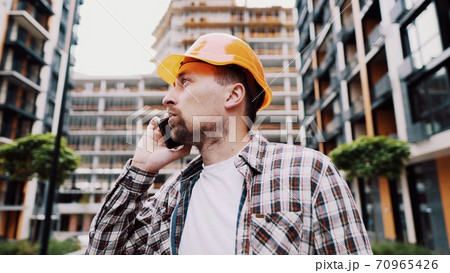 Portrait of young handyman making call while standing at construction area. Engineer talking on the phone on a construction site. Builder in helmet control according to plan by mobile phone Portrait of young handyman making call while standing at construction area. Engineer talking on the phone on a construction site. Builder in helmet control according to plan by mobile phone 70965426