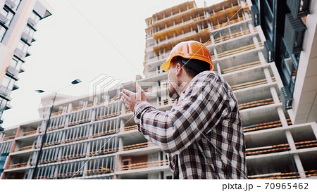 Portrait of young handyman making call while standing at construction area. Engineer talking on the phone on a construction site. Builder in helmet control according to plan by mobile phone Portrait of young handyman making call while standing at construction area. Engineer talking on the phone on a construction site. Builder in helmet control according to plan by mobile phone 70965462