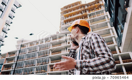 Caucasian male construction worker in orange hard hat and plaid shirt talking on phone at construction site. Architecture theme. Male profession. Foreman controls construction process by smartphone Caucasian male construction worker in orange hard hat and plaid shirt talking on phone at construction site. Architecture theme. Male profession. Foreman controls construction process by smartphone 70965464