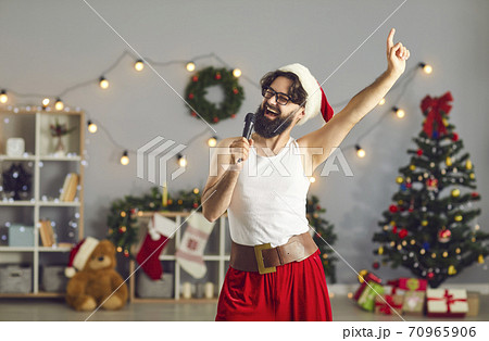 Funny smiling man Santa in festive home clothing celebrating Christmas and singing in microphone at home Funny smiling man Santa in festive home clothing celebrating Christmas and singing in microphone at home 70965906