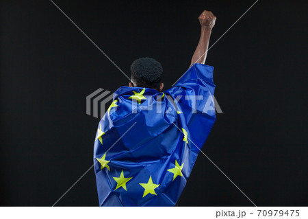 Back view of young african american man wrapped in the flag of the European Union standing with raised fist isolated on black background. Concept of the European Union, unification, integration Back view of young african american man wrapped in the flag of the European Union standing with raised fist isolated on black background. Concept of the European Union, unification, integration 70979475