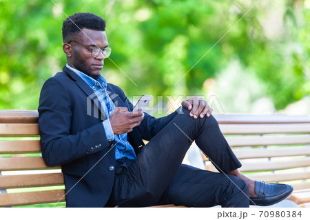 Young African entrepreneur is focused on work issues and communicating on a mobile phone for business deals sitting on a bench in a summer park 70980384