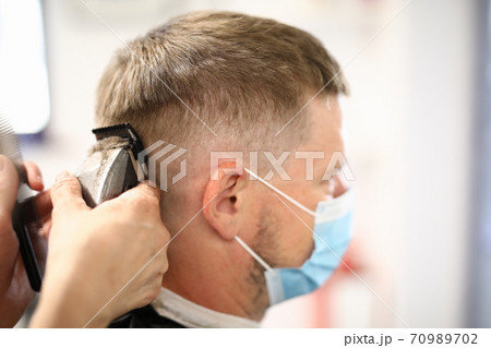 Young man wearing protective face mask gets haircut with clipper in hairdresser 70989702