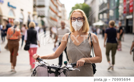 Woman walking by her bicycle on pedestrian city street wearing medical face mask in public to prevent spreading of corona virus. New normal during covid epidemic. Woman walking by her bicycle on pedestrian city street wearing medical face mask in public to prevent spreading of corona virus. New normal during covid epidemic. 70995694