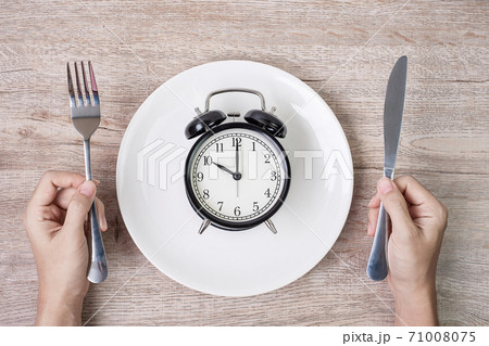 Hands holding knife and fork above alarm clock on white plate on wooden table background. Intermittent fasting, Ketogenic dieting, weight loss, meal plan and healthy food concept 71008075