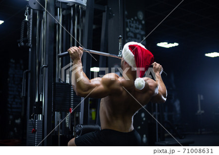 Young sexy Santa Claus in a Christmas hat is working out pumping up back muscles in the gym gaining weight on machines 71008631