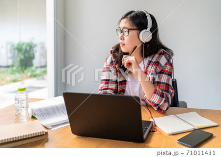Young Asian female student wearing headphone looking out the window while working and studying from home Young Asian female student wearing headphone looking out the window while working and studying from home 71011431