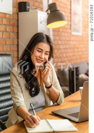Happy young beautiful Asian business woman taking on the phone while using a computer during working from her home office during COVID pandamic lockdown 71016393