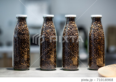 Glass transparent bottles filled with roasted coffee beans, transparent containers with a white lid stand on the table of the store's matrine. Glass transparent bottles filled with roasted coffee beans, transparent containers with a white lid stand on the table of the store's matrine. 71018968
