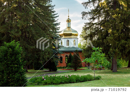 Orthodox church with a golden dome and a cross on its top, a religious building among trees and a landscape with flower beds and flowers. 71019792