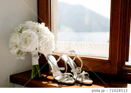 White sandals of the bride on the windowsill with a bouquet of roses on a wooden windowsill by the window overlooking the mountains. White sandals of the bride on the windowsill with a bouquet of roses on a wooden windowsill by the window overlooking the mountains. 71028512