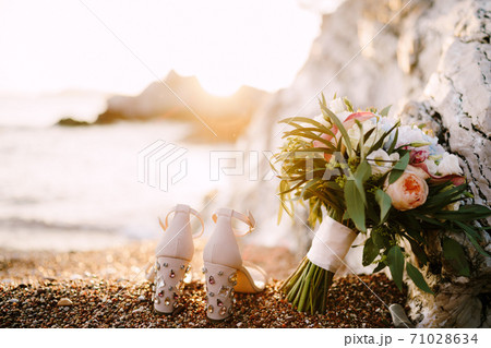Bridal shoes with precious. stones on heels, on the sea pebble beach, bridal bouquet on the background of the sunset. 71028634