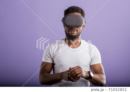 Handsome young African man in VR headset standing against purple background Handsome young African man in VR headset standing against purple background 71032852