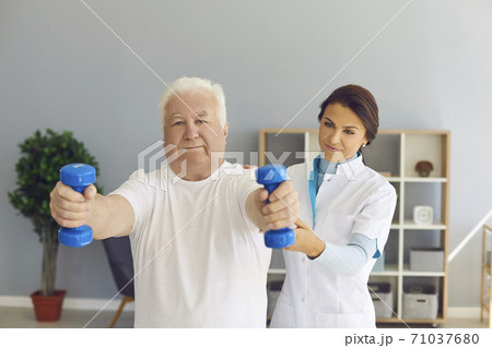 Experienced female doctor supports senior man's arms as he lifts dumbbells in medical office. Experienced female doctor supports senior man's arms as he lifts dumbbells in medical office. 71037680