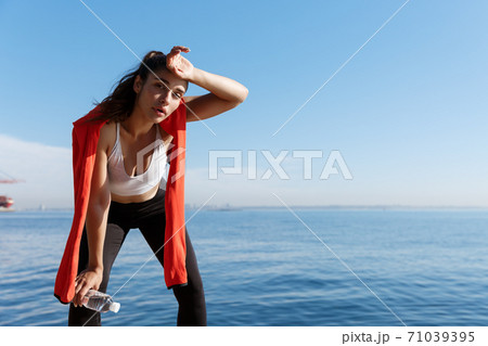 Outdoor shot of tired young sportswoman having a break near the sea, wiping sweat and panting after running 71039395