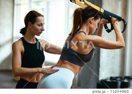 Suspension Training. Young athletic woman in sportswear exercising with trx fitness straps with assistance of personal trainer at gym 71054960
