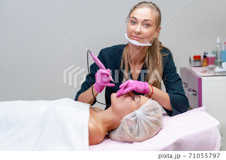 Cosmetology cabinet client lies on couch. Beautician applies marking on eyebrows. Cosmetology cabinet client lies on couch. Beautician applies marking on eyebrows. 71055797