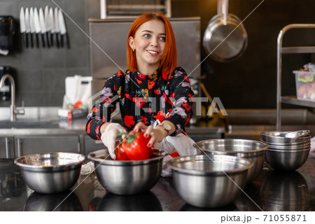 Beautiful young red-haired girl in evening dress is preparing food in a professional kitchen. 71055871