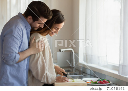 Caring millennial husband hugging shoulders of beloved wife preparing breakfast Caring millennial husband hugging shoulders of beloved wife preparing breakfast 71056141