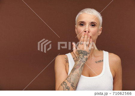 Manlike woman in white vest covering mouth by hand and showing arm with tattooes Manlike woman in white vest covering mouth by hand and showing arm with tattooes 71062566