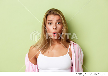 Close-up of attractive blond woman in pink shirt, looking surprised, saying wow fascinated, standing over green background 71074527