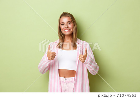 Portrait of beautiful woman with perfect white smile, showing her support and approval, make thumbs-up, standing over green background 71074563