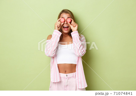Portrait of silly blond girl playing with candies, holding macaroons over eyes and looking amazed, standing against green background 71074841