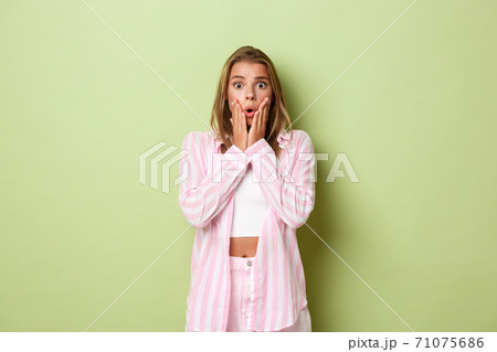 Portrait of shocked blond girl in pink outfit, gasping and looking nervous, standing alarmed against green background 71075686