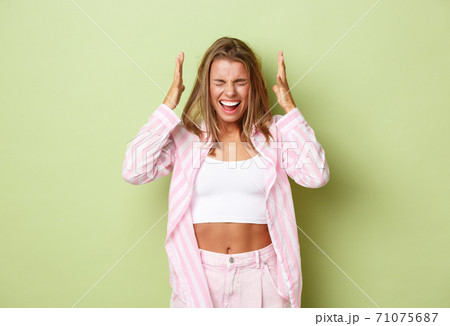 Potrait of distressed blond woman in pink shirt, holding hands on head and screaming, having depression, standing over green background 71075687