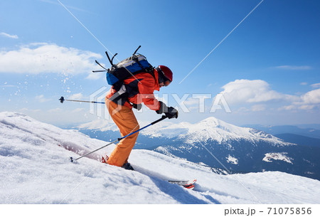 Sportsman skier in bright clothing, helmet and goggles with backpack riding down steep snowy slope. 71075856