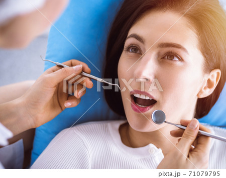 Smiling brunette woman being examined by dentist at sunny dental clinic. Hands of a doctor holding dental instruments near patient's mouth. Healthy teeth and medicine concept 71079795