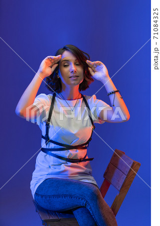 Young dark-haired woman with short hair sits on chair with hands up straightening her hair. Cheerful Caucasian lady isolated on blue background. Young dark-haired woman with short hair sits on chair with hands up straightening her hair. Cheerful Caucasian lady isolated on blue background. 71084325