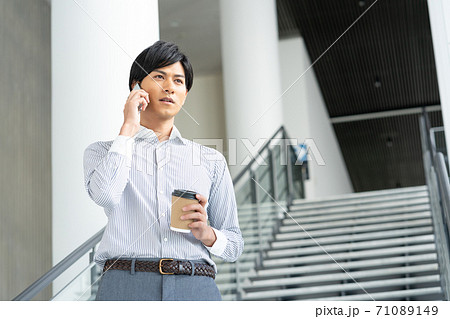 オフィスのロビーで飲み物を飲みながらスマホで会話をする若い男性 71089149