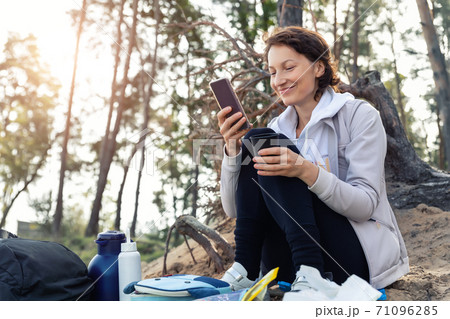Young adult caucasian beautiful attractive female person enjoy having fun use phone and drink warm drink at picnic hiking trip pine forest wood autumn day. Family nature outdoor recreation lifestyle 71096285