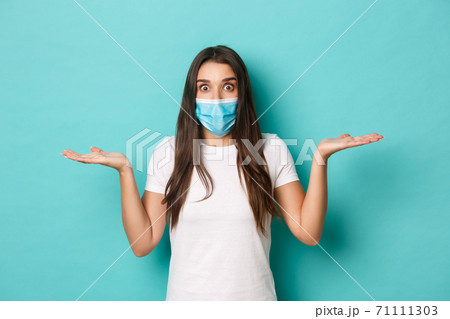 Concept of pandemic, covid-19 and social distancing. Image of clueless young woman in white t-shirt and medical mask, looking amazed and shrugging, standing over blue background 71111303