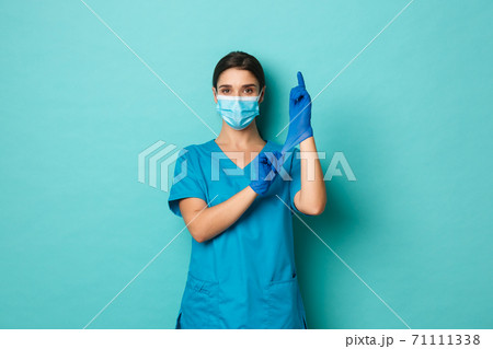 Covid-19, pandemic and medicine concept. Confident beautiful woman in medical mask and crubs, put on gloves and looking self-assured, getting ready for patient check up Covid-19, pandemic and medicine concept. Confident beautiful woman in medical mask and crubs, put on gloves and looking self-assured, getting ready for patient check up 71111338