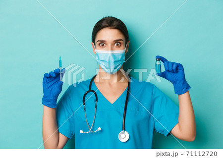 Concept of covid-19 and quarantine concept. Close-up of female doctor in medical mask, latex gloves and scrubs holding syringe and showing coronavirus vaccine, looking amazed 71112720