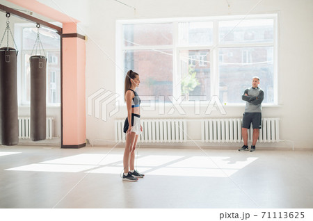 In a bright gym with large windows a young female athlete jumps on a rope under the supervision of a coach 71113625