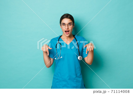Coronavirus, pandemic and social distancing concept. Image of attractive female doctor in scrubs, pointing fingers down and smiling fascinated, showing logo, standing over blue background 71114008