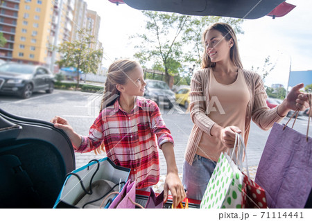 Girl and young female looking at each other, taking out shopping bags from car trunk 71114441