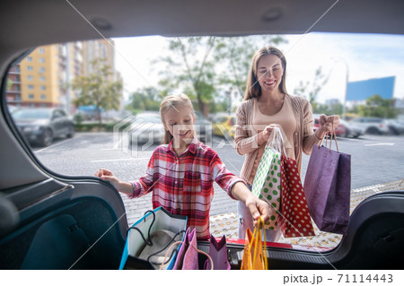 Fair-haired girl and young female taking out shopping bags from car trunk 71114443